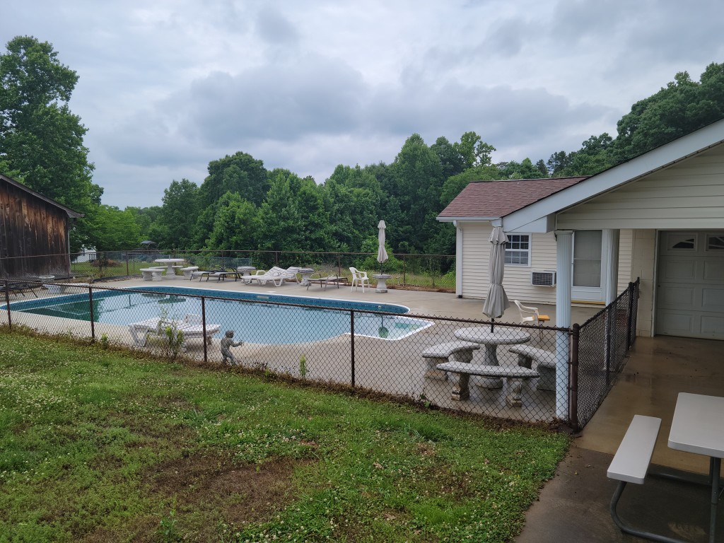 In-ground swimming pool with concrete patio, lounge chairs, and stone table in a fenced backyard